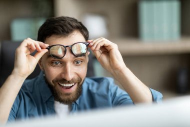 Heyecanlı genç iş adamı gözlüğünü çıkarıp bilgisayar ekranına bakarken, Mutlu Milenyum Erkeğinin Çevrimiçi Haberlere Duygusal Tepkisi, Seçici Odaklanma