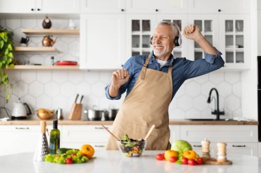 Mutfakta yemek pişirirken müzik dinleyen, sağlıklı yemek hazırlayan, dans eden, evde öğle yemeği yaparken eğlenen neşeli, yaşlı ve yaşlı bir adam.