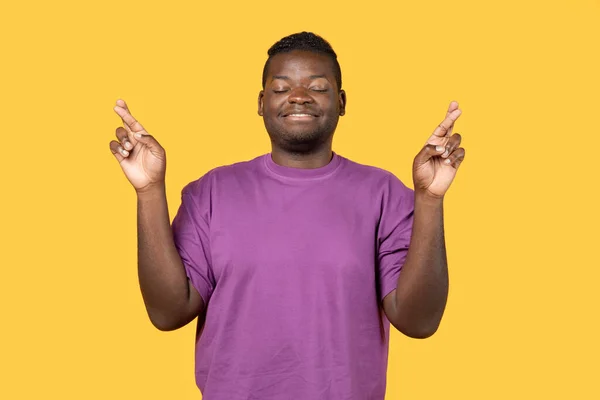 希望的好运 年轻的黑人小伙子闭眼许愿 祈求奇迹站在黄色工作室的背景上 穿着便衣紫色T恤衫 — 图库照片