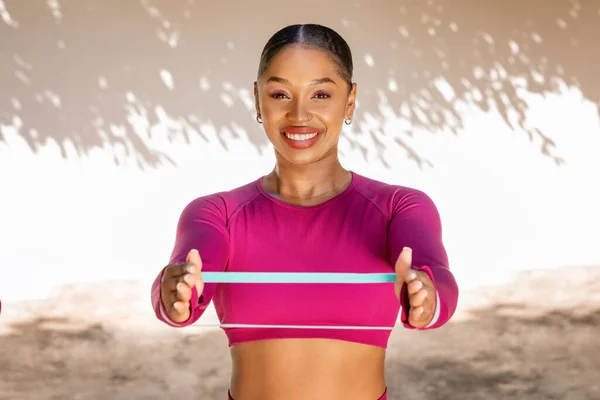 Athletic african american sportswoman exercising with stretch band outdoor. Happy young black lady in pink top holding resistance band, working on her arms strength. Fitness tools, accessories