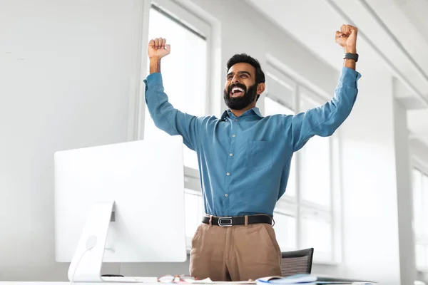 Joyful indian businessman celebrating success in front of computer at workplace in office, shaking fists and shouting Yes, emotionally reacting to good news. Big luck celebration concept