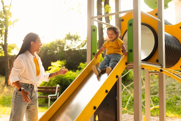 Playground Adventures. Excited Chinese Toddler Infant Girl Giggling as She Rides Down the Slides And Mommy Catching Her at Amusement Park Outside. Babyhood Joy and Playtime Fun