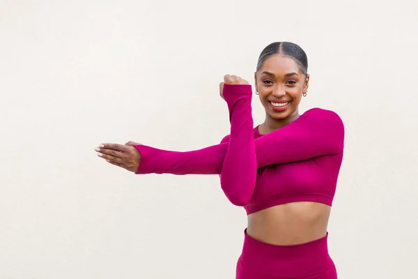 Sporty young african american woman in pink sportswear posing on white blank wall background, stretching arms, copy space for advertisement, sportswoman demonstrating perfect body curves