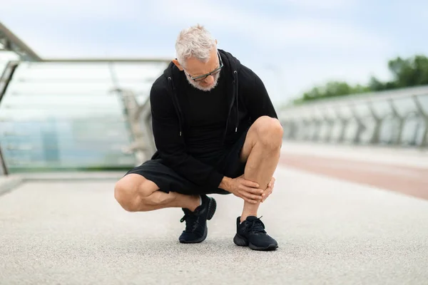 Senior man wearing black sportswear jogging outdoor by bridge, squating and touching his leg, retired sportsman suffering from shin splint, got injured during workout, copy space