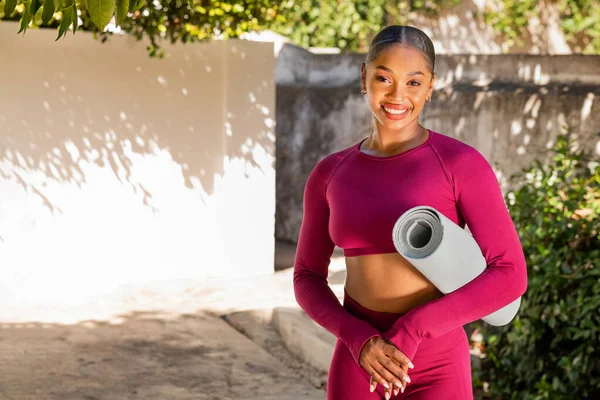 Beautiful positive young black woman in pink tight sportswear have outdoor workout at garden, sportswoman holding fitness mat and smiling at camera, practicing pilates, yoga, stretching, copy space