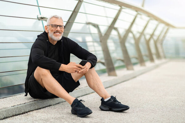 Happy grey-haired retired sportsman in black sportswear sitting on ground, using smartphone, have rest while exercising outdoor jogging by bridge, track workout progress on fitness app, blank space