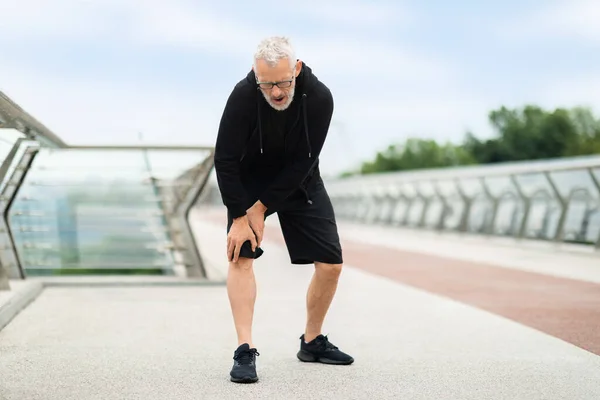 Senior man runner suffering with knee pain during workout outdoor. Elderly sportsman running by footbridge, touching his leg, have muscle strain, copy space. Exercise and injury prevention in seniors