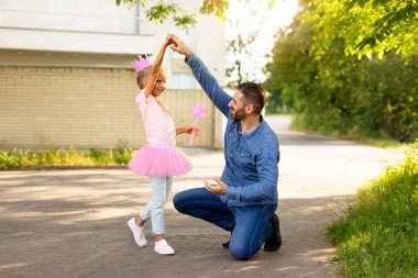 Sevgi dolu baba tek dizinin üzerinde duruyor, kollarını kaldırıyor, küçük kızını eğiyor, parkta yürürken prenses gibi giyiniyor. Aile müzikle dans ediyor, birlikte eğleniyor.