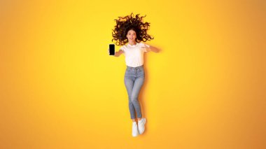 Mobil Uygulama Reklamı. Neşeli Kadın Cep telefonunu işaret ediyor, Yeni Gadget 'ı gösteriyor ve Dijital Teklif' i tanıtıyor, Sarı Stüdyo Arkaplanı Üzerinde Kameraya Gülümseyen İnternet sörfü yapıyor. Panorama, Tam Uzunluk