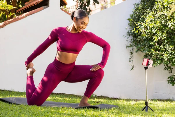 Positive well-fit young black woman in pink tight sportswear exercising on yoga mat at garden, watching online fitness class on smartphone set on tripod, enjoying morning pilates workout