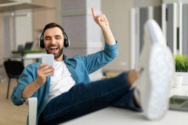 Neşeli genç ofis çalışanı kulaklıkla müzik dinliyor ve işyerinde akıllı telefon kullanıyor, Milenyum 'lu iş adamı masanın üstünde bacaklarıyla dinleniyor, işten ayrılmanın keyfini çıkarıyor,
