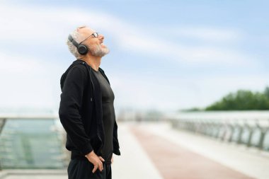 Neşeli sporcu kıdemli sporcu, dışarıda antrenman yaparken, köprüde, gözleri ve elleri belinde, kablosuz kulaklıkla müzik dinlerken, fotokopi çekerken,