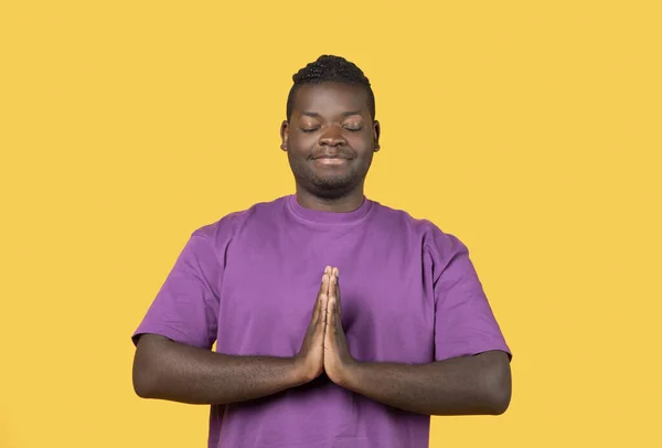身穿紫色T恤的安静的黑人年轻人手挽手靠近胸部 闭上眼睛祈祷 在黄色的背景上做着祈祷手势 工作室拍摄的家伙在祈祷 — 图库照片