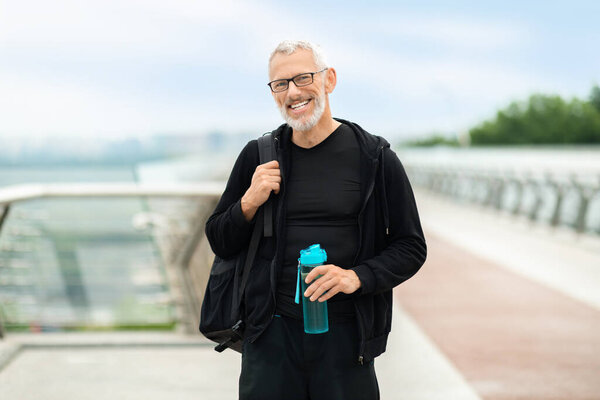 Happy senior sportsman drinking water after workout outdoor. Cheerful grey-haired man in sportswear with backpack holding bottle of water, smiling at camera, stand on bridge, going home afer jogging