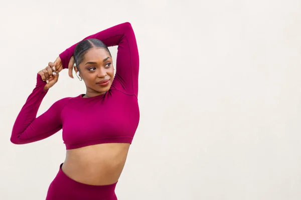 Sexy athletic young black woman in pink sportswear posing on white blank wall background, looking at copy space for advertisement, sportswoman demonstrating perfect body curves, using earpods