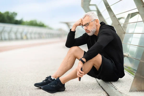 Exhausted elderly sportsman sitting on ground, touching his head, holding smartphone, retired man working out outdoor, jogging by bridge, feeling tired after running, copy space