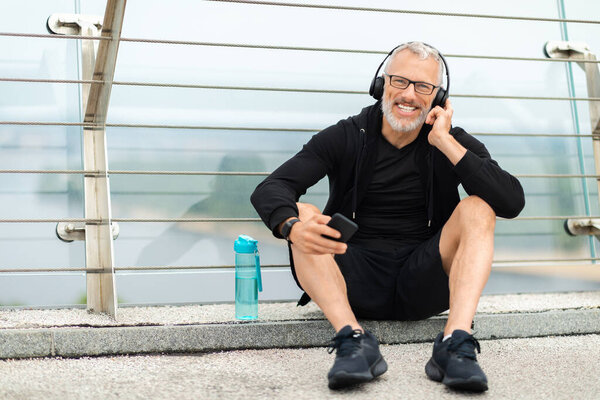 Happy senior sportsman sitting on the ground, drink water after workout outdoor, touch button on wireless headphones, hold smartphone, elderly man answer call while exercising on street, copy space