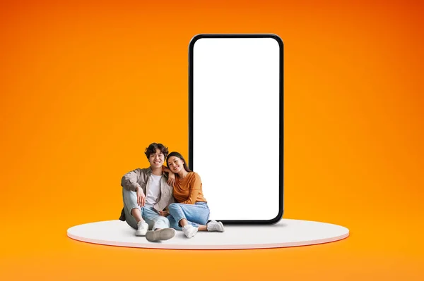 Nice online offer, great deal for two. Smiling young korean couple man and woman friends sitting by big phone over orange backgorund, embracing, white blank screen, blank space, mockup