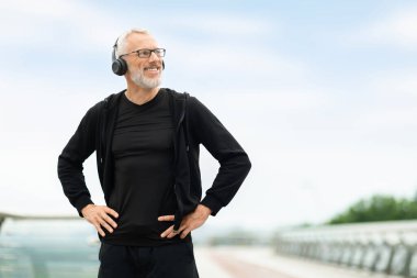 Neşeli gri saçlı yakışıklı büyükbaba siyah spor giysi ve kablosuz kulaklık takıyor. Sabah antrenmanlarına dışarıda çıkıyor. Emekli sporcu koşudan sonra dinleniyor.