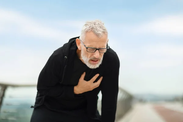 Sport and cardiovascular diseases, physical activity and injuries in seniors. Closeup of retired sportsman in black sportswear touching chest, have heart pain during outdoor workout, copy space