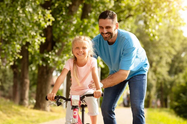 Loving european middle aged father and his pretty daughter spending time together oudoors, dad teaching child how to ride a bicycle, walking in park