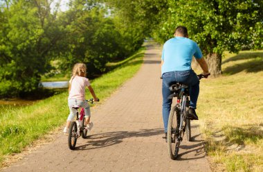 Orta yaşlı bir baba ve küçük kızının parkta bisiklet sürerken, hafta sonları yürüyüş yaparken ve birlikte vakit geçirirken, güneşli havanın tadını çıkarırlarken görüntüsü. Fiziksel aktivite, eğlence.