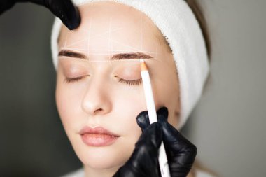 Professional Cosmetologist Mapping Eyebrows Of Young Woman With White Pencil Before Permanent Makeup Treatment In Beauty Salon, Marking Up Brow Zone And Forehead Before PMU Procedure, Closeup Shot clipart