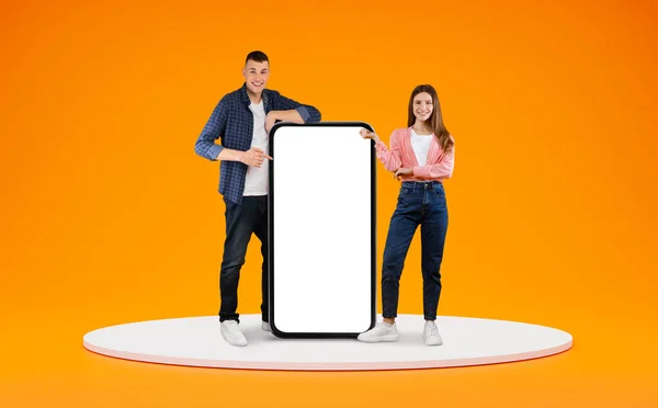 Mobile Offer. Happy Young Couple Pointing At Big Cellphone With White Screen For Mockup, Cheerful Man And Woman Demonstrating Copy Space For Phone Application Design, Posing Over Orange Background