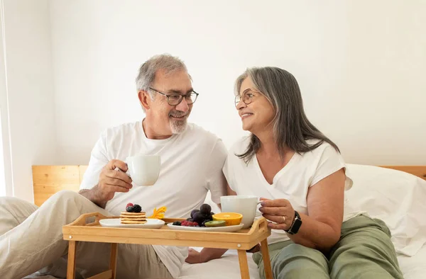 Happy old european husband and wife have breakfast, enjoy coffee cups, sit on bed in bedroom interior. Rest and relaxation together, healthy food at home, romantic relationships