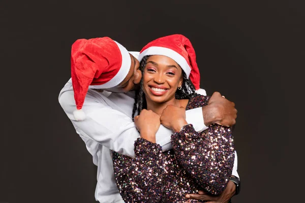 Portrait of happy millennial black couple wearing nice outfits celebrating Christmas together on black background. Loving african american man kissing and hugging his smiling beautiful wife