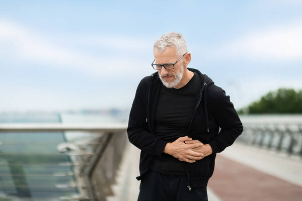 Senior sportsman exercising outdoor, standing on bridge, touching left side, experiencing acute pain while having workout on street, copy space. Abdominal pain and sports concept