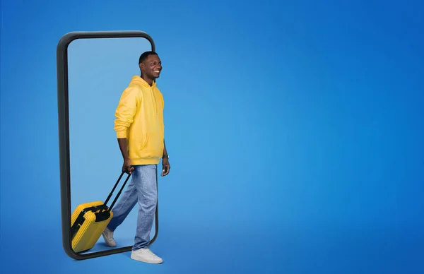 穿着运动衫的年轻的非洲裔美国人 拿着手提箱从带有空白屏幕的大智能手机中走出来 在蓝色的工作室背景下被隔离 带装置休假 周末的应用程序 — 图库照片