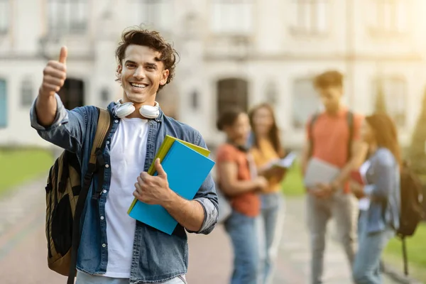 International Study Programs. Handsome Young Male Student Showing Thumb Up At Camera While Posing Outdoors At University Campus, Happy Guy Holding Workbooks, Recommending Education Abroad