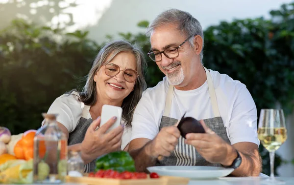 Cheerful old european couple in aprons with glasses of wine use smartphones, chatting in garden, outdoors. Lunch holiday together, device for video call, technology and fun