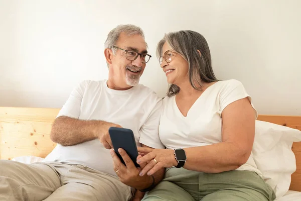 Happy old european husband and wife watch video on phone, chatting, sit on bed in bedroom interior. Rest and relaxation together, photo app and device, fun at spare time at home