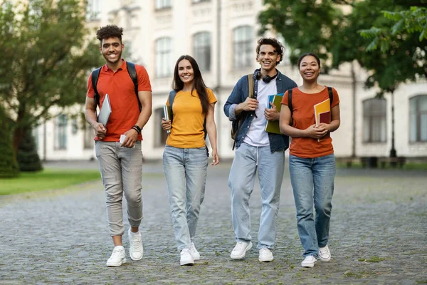 Portrait Of Happy Cheerful College Friends Walking Outdoors After Classes, Positive Young Multiethnic Students Carrying Workbooks And Backpacks, Smiling And Looking At Camera While Going To Campus