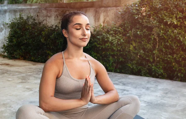 Calm happy young arab woman athlete in sportswear with closed eyes practice yoga, meditate in lotus position, enjoy breathing exercises, outdoor at summer morning. Sports, fit, workout alone