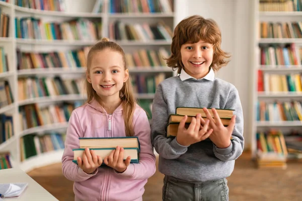 一个快乐的欧洲男孩和女孩拿着一堆书 站在图书馆里 对着相机微笑的画像 孩子们手里拿着一堆书 — 图库照片