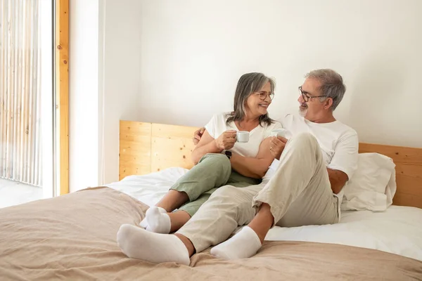 Cheerful old european man and lady have breakfast with cups of coffee, enjoy free time, sit on bed in bedroom interior. Communication and relationships in morning at home, rest and relax together