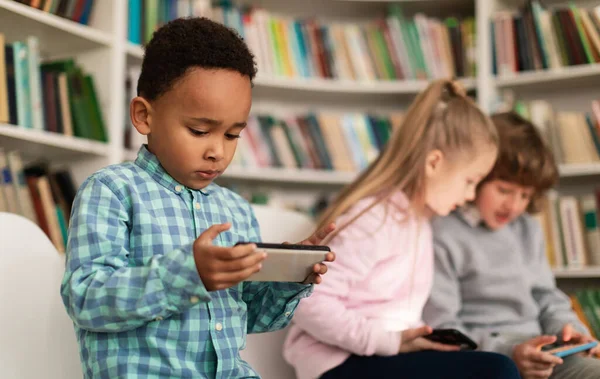 School children playing video games on smartphones during break while sitting in library or classroom, selective focus on african american boy. Modern school concept
