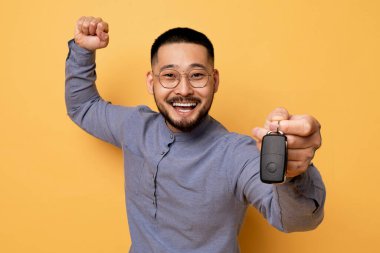 Komik Asyalı genç adam, kamerada araba anahtarını gösteriyor, Milenyum 'da yeni araba kiralama ya da araba kiralama servisini kutlayan neşeli heyecanlı adam, stüdyoda sarı arka planda duruyor,