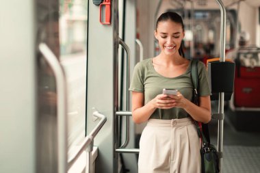 Modern Tramvayda ayakta cep telefonuyla mesajlaşan neşeli Yolcu Kadın. Gezgin Bayan İşe Yeni Akıllı Telefonla Gidiyor. Modern Ulaşım Tatil, Şehir Seyahati ve İletişim