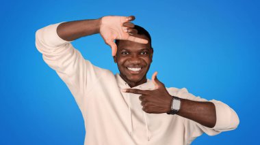 Sweatshirt 'lü neşeli Afro-Amerikalı adam mavi stüdyo arka planında, el çerçevesi ya da lensli işaret dilini kullanıyor. Pozitif yaşam tarzı, yaratıcı sanat, fotoğraf, reklam ve teklif
