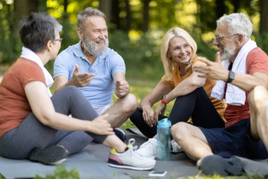 Sportif son sınıf öğrencileri spor antrenmanından sonra birlikte dinleniyor, neşeli olgun erkek ve kadınlar dışarıda yoga minderlerinde oturuyor ve sohbet ediyor, spor eğitiminden sonra dinleniyor, yakınlaşıyorlar.