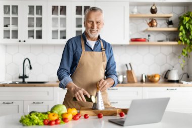 Olgun Şef. Mutfakta Önlüklü Kıdemli Adam Yemek Pişiriyor, Yakışıklı Yaşlı Adam Evde Sağlıklı Sebze Yemeği hazırlarken Dizüstü bilgisayar kullanıyor, Kabak rendeliyor ve kameraya bakıyor