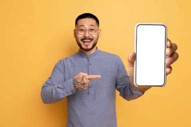 Şuna bak. Neşeli Asyalı adam elinde beyaz beyaz ekranla koca akıllı telefonu işaret ediyor, mobil uygulama ya da web sitesi tasarımı için bedava kopya alanı gösteriyor, sarı arka planda duruyor,
