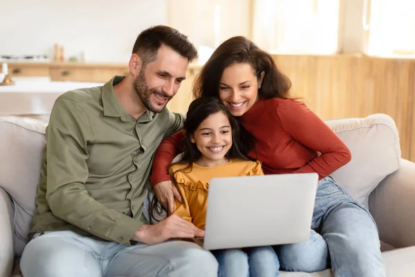 Online Offer. Cheerful Parents And Daughter Using Laptop, Websurfing And Typing, Family Spending Time Together With Computer, Visiting Internet Website At Home, Sitting On Sofa Indoor
