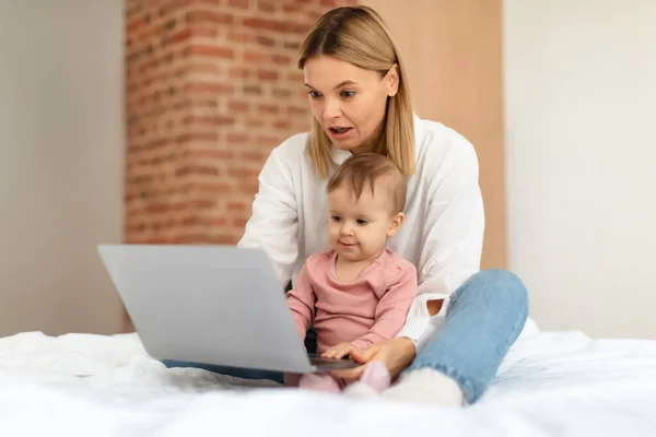 Baby and gadgets. Mommy and her little child daughter using laptop, browsing internet for online fun together, sitting on bed at home, free space. Mom and infant kid watching cartoons on computer