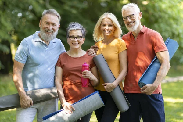 Group of happy senior people practicing sport together outdoors, smiling mature men and women holding fitness mats and looking at camera, elderly friends enjoying active lifestyle on retirement
