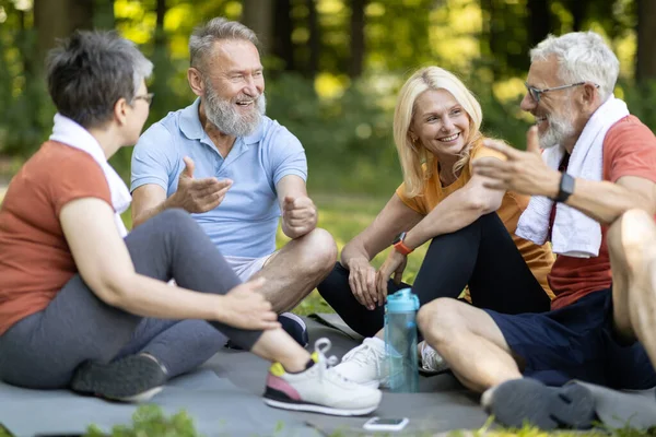 Group of sporty senior people resting together after outdoor fitness workout, cheerful mature men and women sitting on yoga mats outside and chatting, relaxing after sport training, closeup